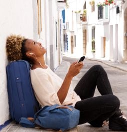 A woman sits on the sidewalk with her bags listening to music on her phone in a small Mediterranean-looking street